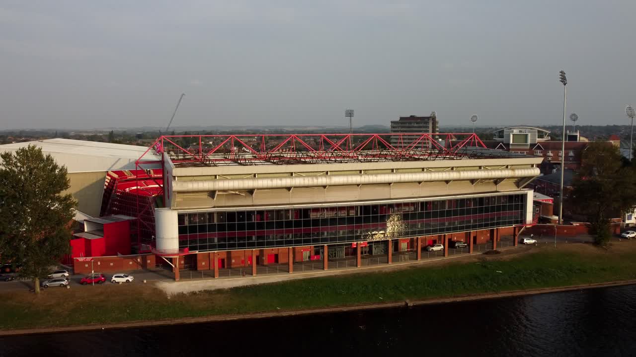 Drone video captures the outside of Nottingham Forest Football Stadium next to the River Trent. The video rotates around one side of the stadium and captures the sun reflecting off the glass.