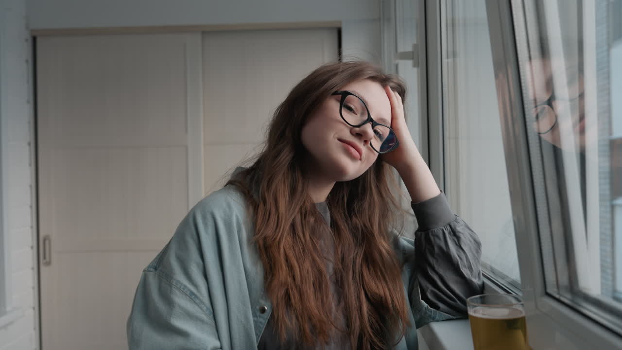 Donna rilassata che sorride vicino alla finestra. Donna soddisfatta che riposa serenamente accanto a una finestra illuminata dal sole, con gli occhiali. Donna tranquilla in abiti comodi che guarda fuori dalla finestra con un sorriso gentile dopo un successo