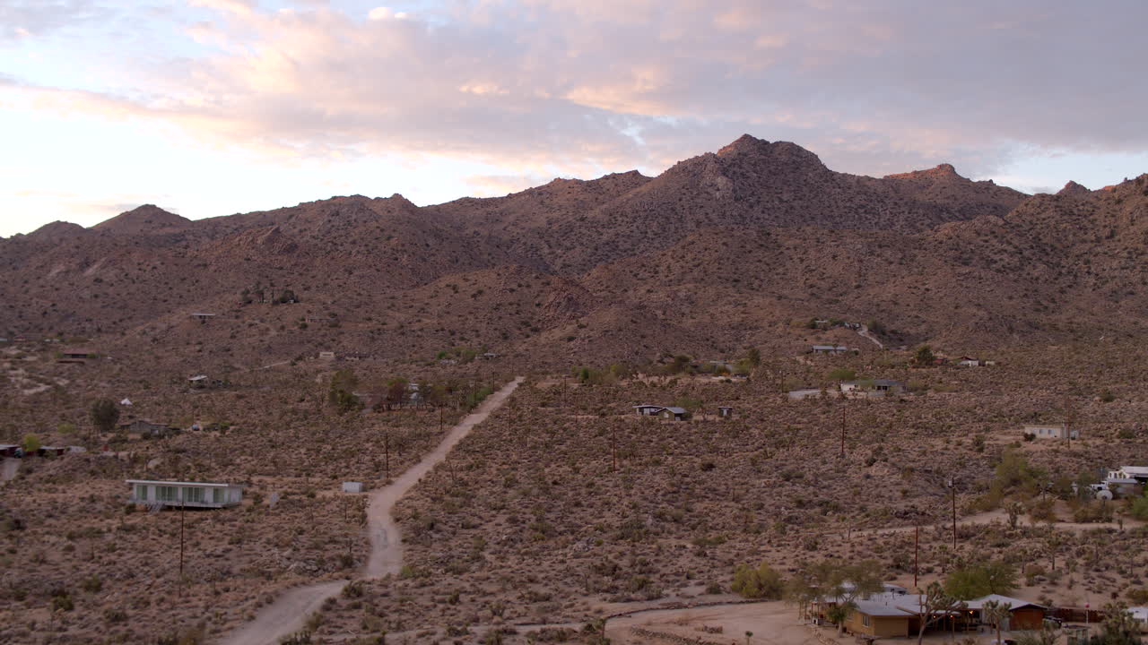 Sorvolo di una strada sterrata nel deserto sopra le case e verso colline frastagliate a Joshua Tree, California, all'alba