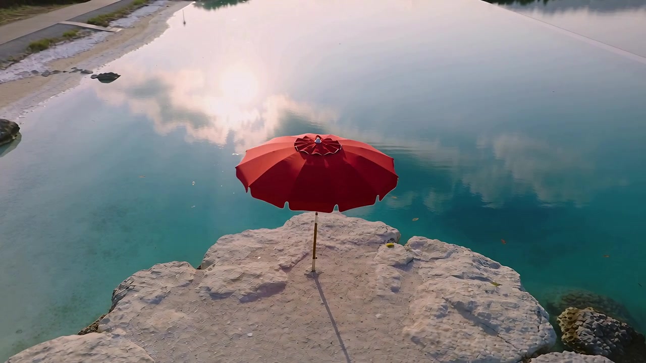 Drone che si allontana per mostrare un ombrellone rosso su una roccia chiara vicino all'acqua turchese, con pedoni su un sentiero