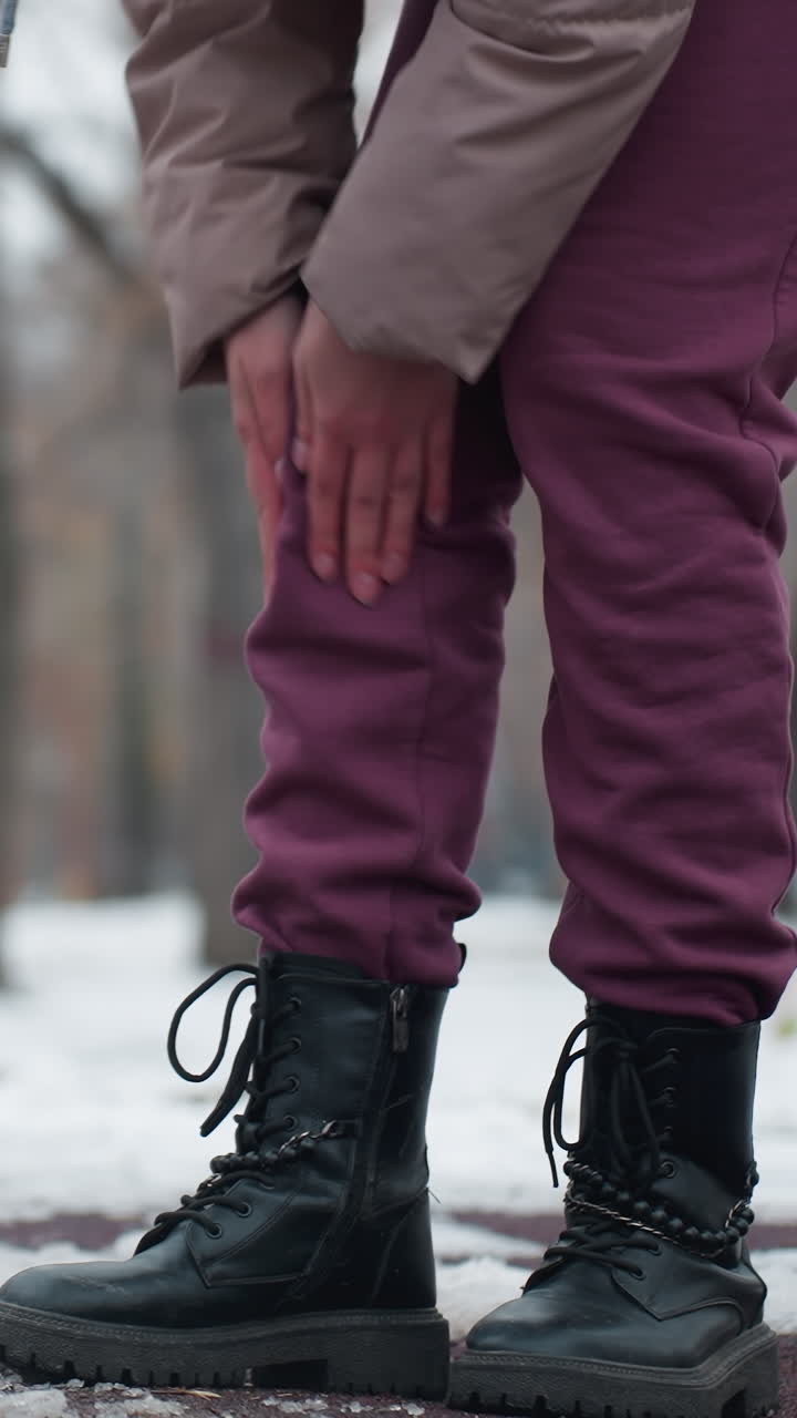 joven con traje de invierno siente dolor en la rodilla, se inclina y se frota la rodilla cerca del columpio en un entorno al aire libre nevado con árboles en el fondo, mostrando incomodidad en el ambiente frío del parque de invierno