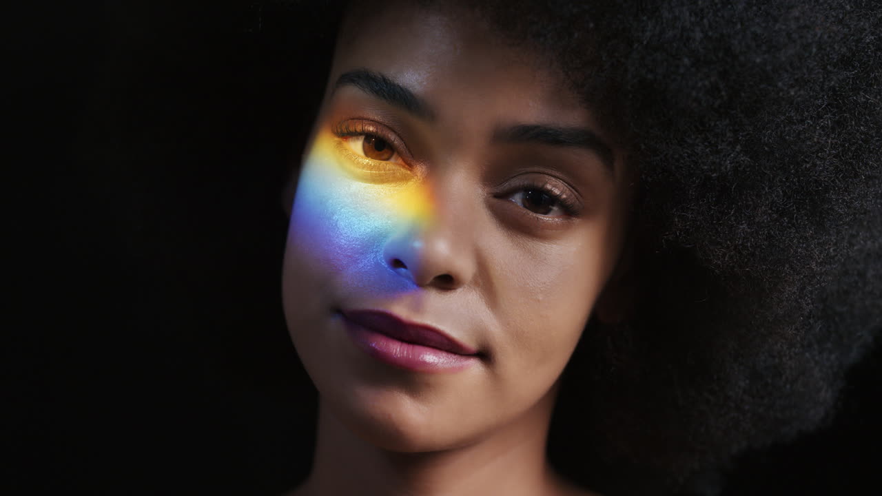 retrato de arco iris de cerca mujer afroamericana atractiva con afro disfrutando de una piel suave y saludable con aspecto confiado belleza natural luz multicolor sobre fondo negro concepto de cuidado de la piel
