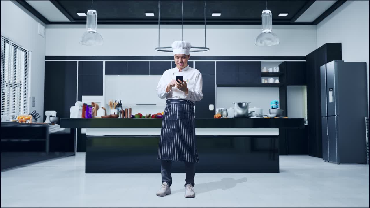 cuerpo lleno de hombre asiático chef sonriendo y usando teléfono inteligente mientras está de pie en la cocina de casa
