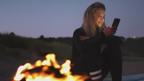 woman sitting on surfboard by camp fire on beach using mobile phone as sun sets behind her