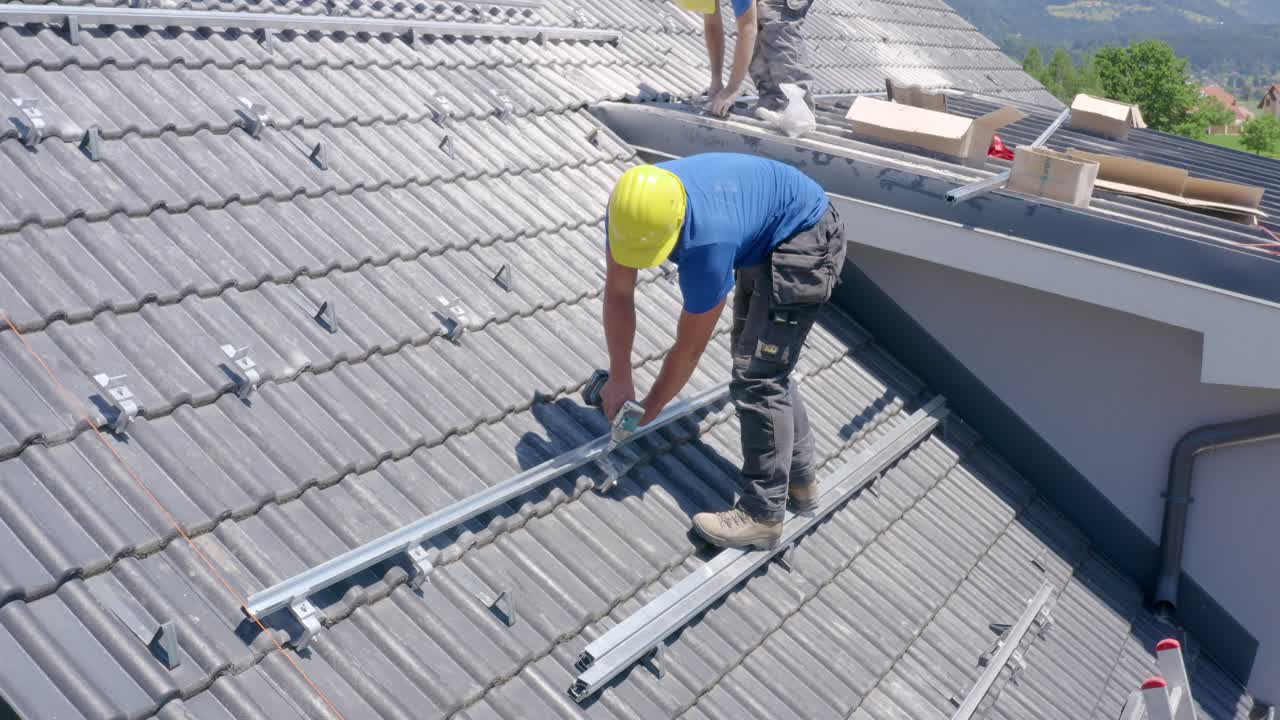 Young Caucasian male solar panel roof installer wearing yellow hard hat standing on steep roof attaches roof anchor with electric portable hand drill, static close up