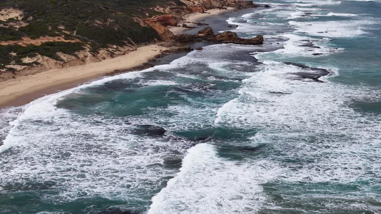Drone captures turquoise waves crashing on rocky shoreline under overcast skies, steady descending movement