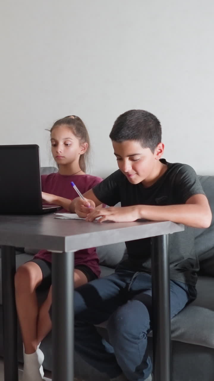 Hermanos haciendo los deberes sentados en el sofá del salón: un niño escribe en un papel y una niña teclea en un portátil, concentrados durante la sesión de estudio vespertina.