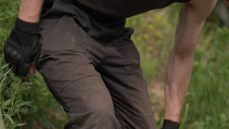 close-up waist view of soldier brushing dirt off gloves and cargo pants after crawling, with muscular arms exposed in sleeveless top, surrounded by natural background and subtle motion of recovery