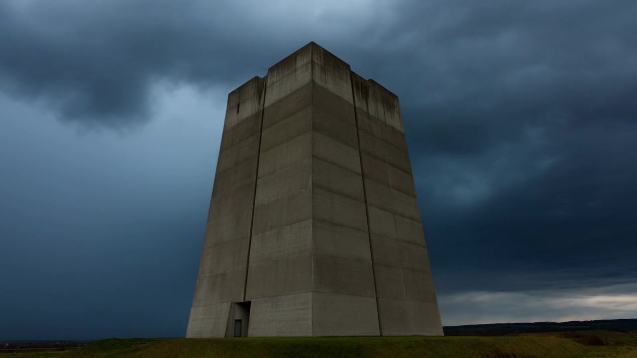 Skiftande stormmoln förmörkar himlen över en gräsbevuxen höjd, vilket förändrar kontrasten och ingången till ett betongtorn.