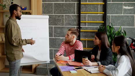 joven empresario afroamericano explicando el plan de negocios de su nueva empresa en flipchart con presentación de tecnología blockchain a sus colegas en una moderna oficina de loft en interiores