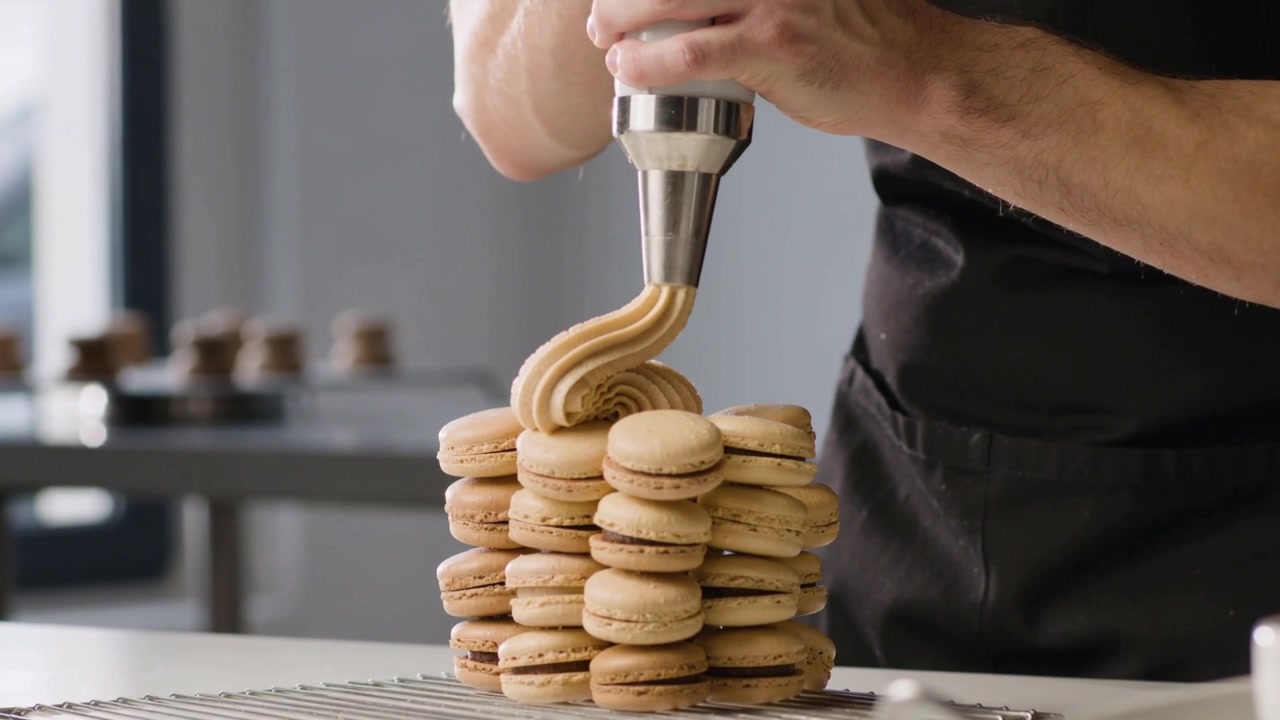 Piping aproned chef entering at counter, pressing metal-tip, icing beige macaron stack for service