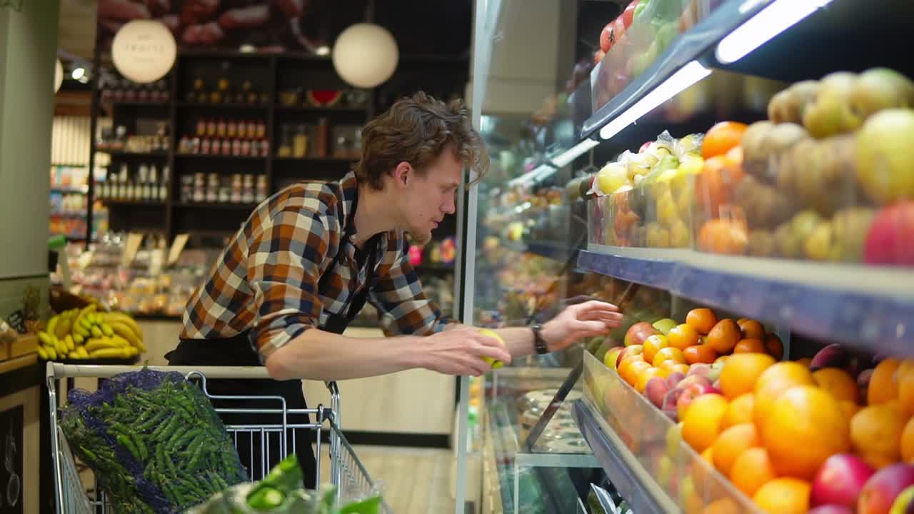 en el supermercado: guapo empleado de almacén con un delantal negro, organizando frutas y verduras orgánicas. agregando manzanas frescas en