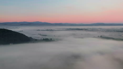 slow, aerial view of town in valley covered by mist with mountain view at sunrise