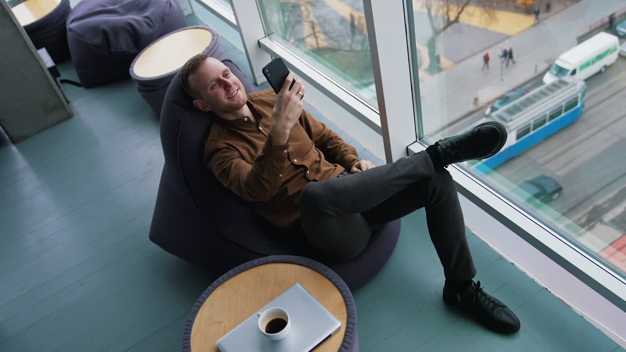 Young businessman sitting at break time. Smiling man sitting by the window and looking into the phone on the city background through the window. Top view.