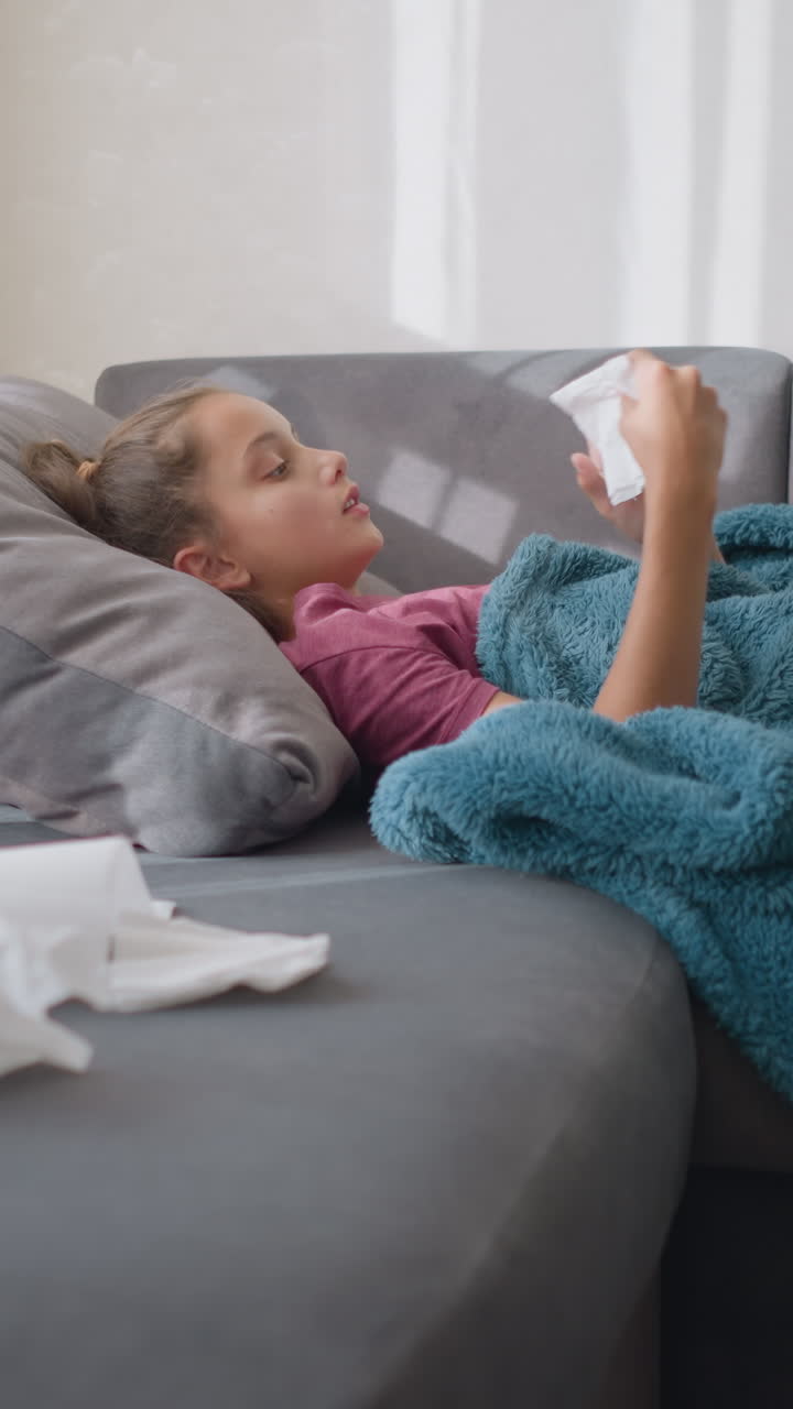 Ragazza con l'influenza sdraiata sul divano, che starnutisce in un fazzoletto di carta con fazzoletti sparsi intorno, con un aspetto sofferente, ambiente accogliente con tazza da tè e fazzoletti visibili sul tavolo, che soffre di malattia