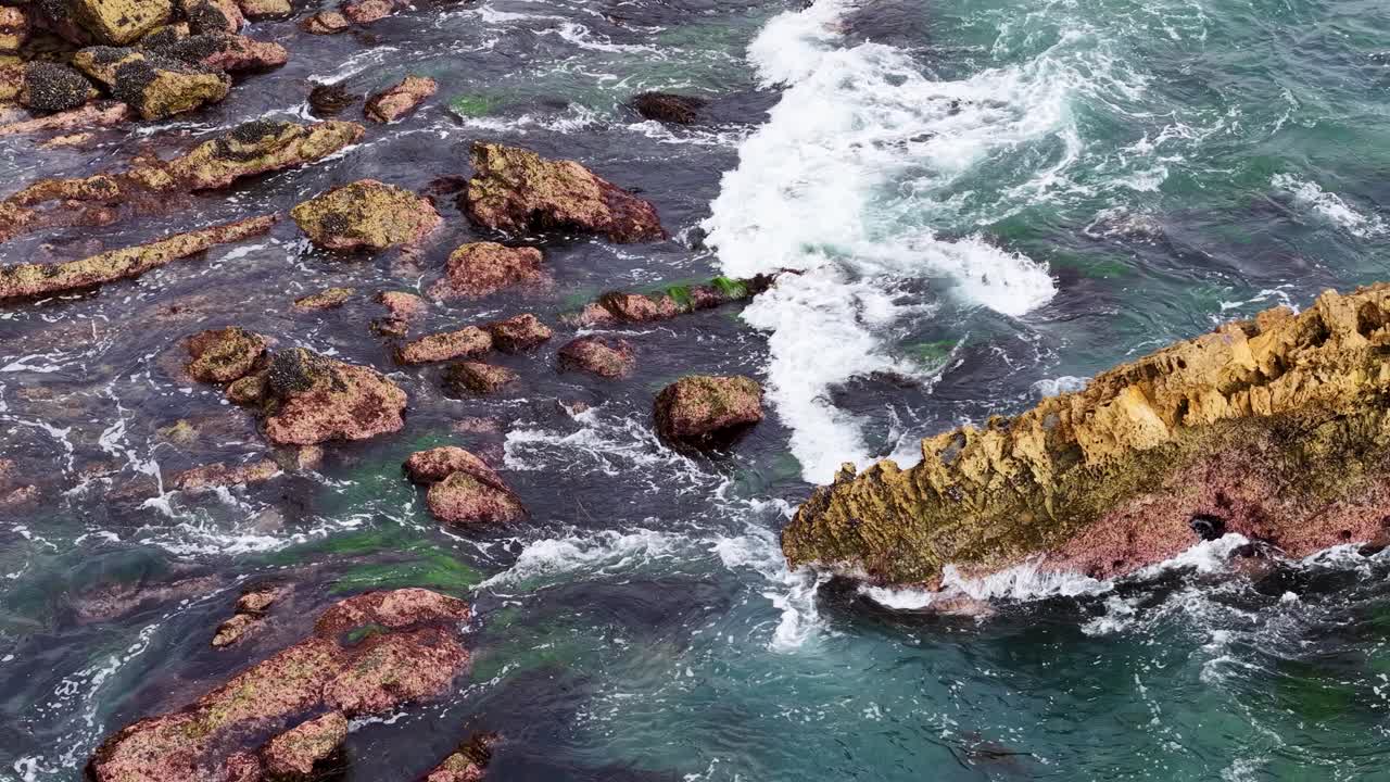 Aerial drone view of rocky coastline as waves crash dramatically against yellow- and red-hued outcroppings