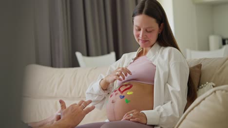 Side-view-of-a-happy-brunette-pregnant-woman-drawing-hearts-on-her-belly-using-bright-colors-together-with-her-husband-while-expecting-a-baby-at-home