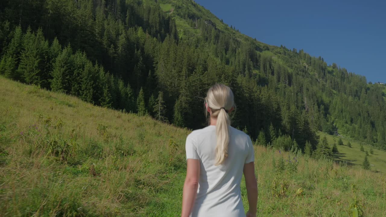 A woman hikes through a green mountain meadow under a clear blue sky on a sunny day