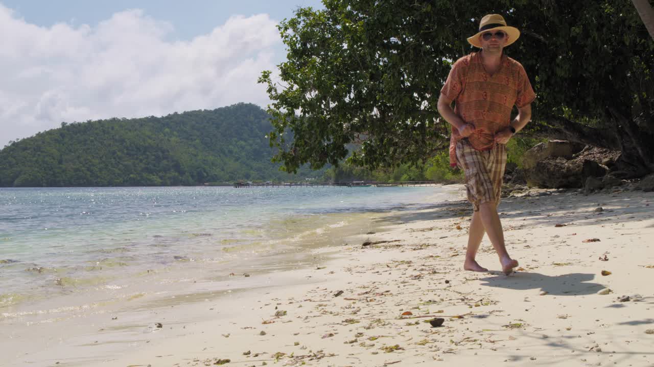 A man walks along the sandy beach of Kri Island, Raja Ampat, Indonesia, enjoying the tropical surroundings. The static shot captures the relaxed pace of island life, with calm waters and lush hills.