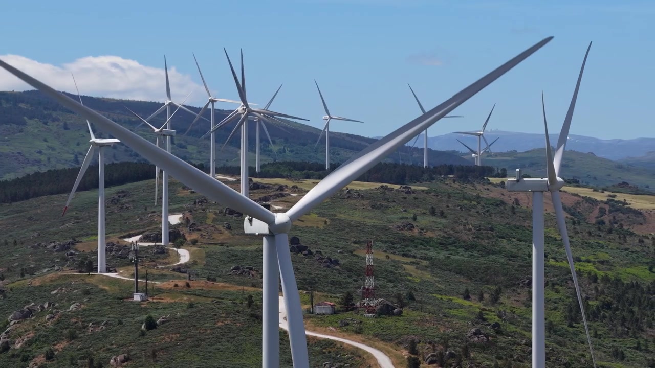 Filmagem aérea de turbinas eólicas modernas girando no topo de um parque eólico montanhoso em Portugal sob céu azul claro. Capturado por drone, este vídeo cinematográfico mostra a energia renovável em ação.