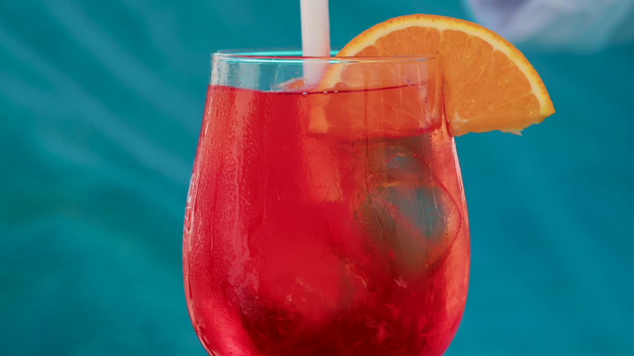 A hand stirs a bright red orange with an orange slice against a turquoise background