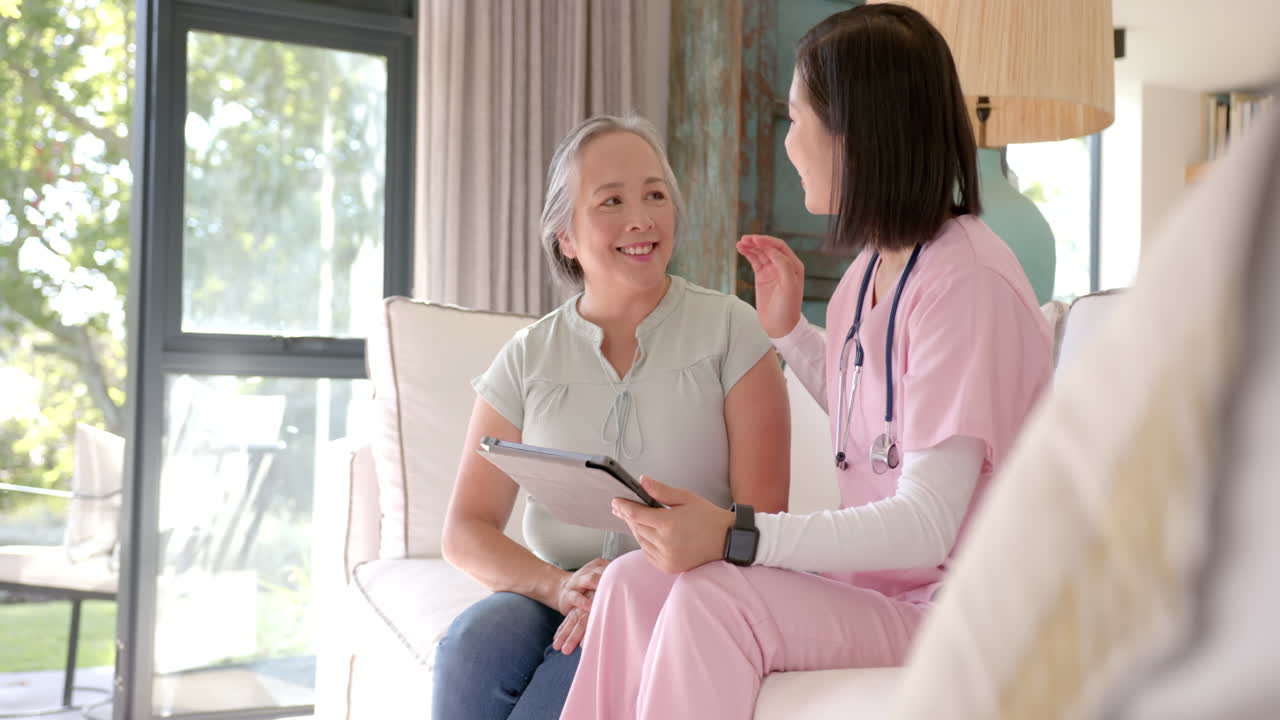 Nurse in pink scrubs consulting senior asian woman at home, holding clipboard and smiling
