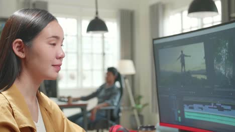 asian female video editor works with footage and sound on his personal computer. she turns and warmly smiles into the camera