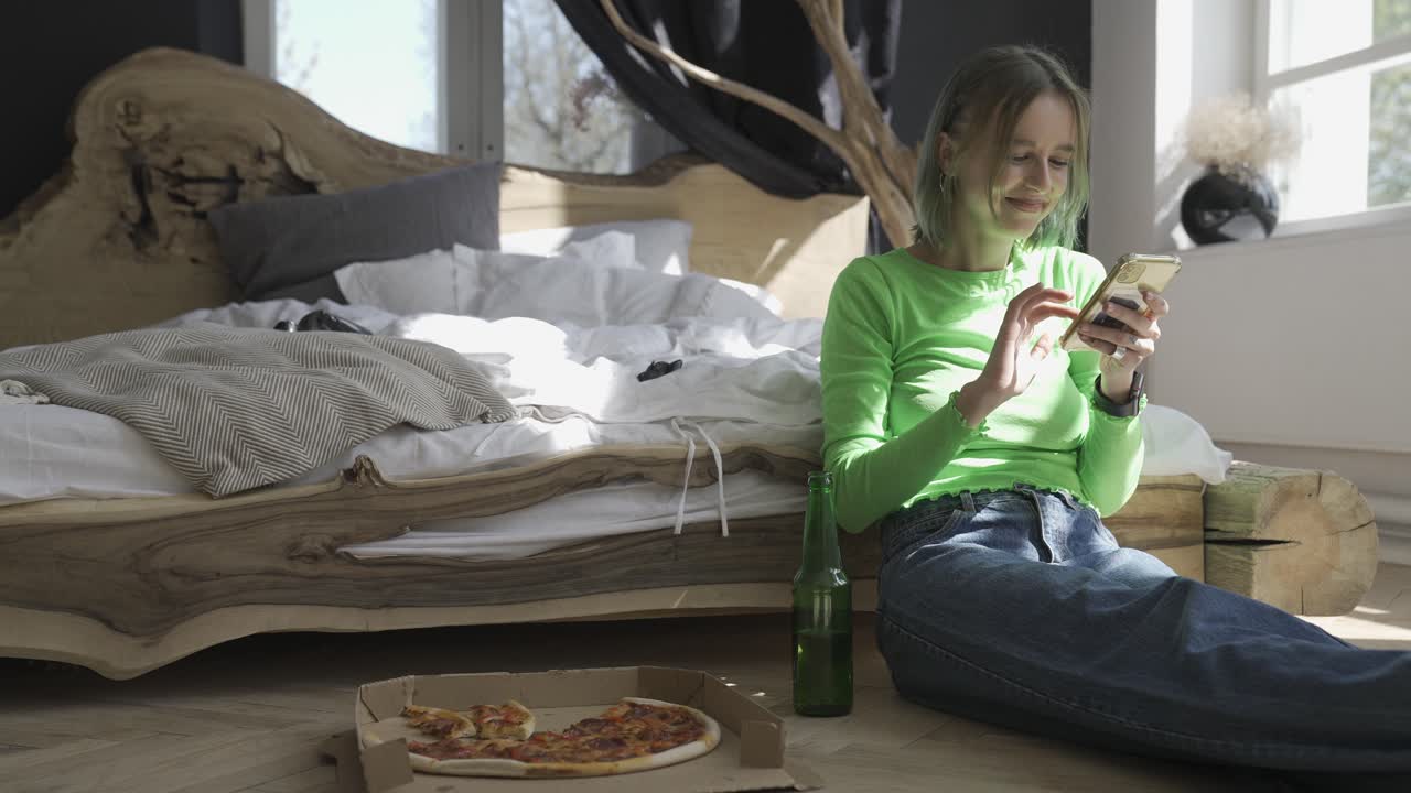 집에서 침실 바닥에 앉아있는 동안 휴대 전화를 사용하고 피자를 먹는 젊은 금발의 여자