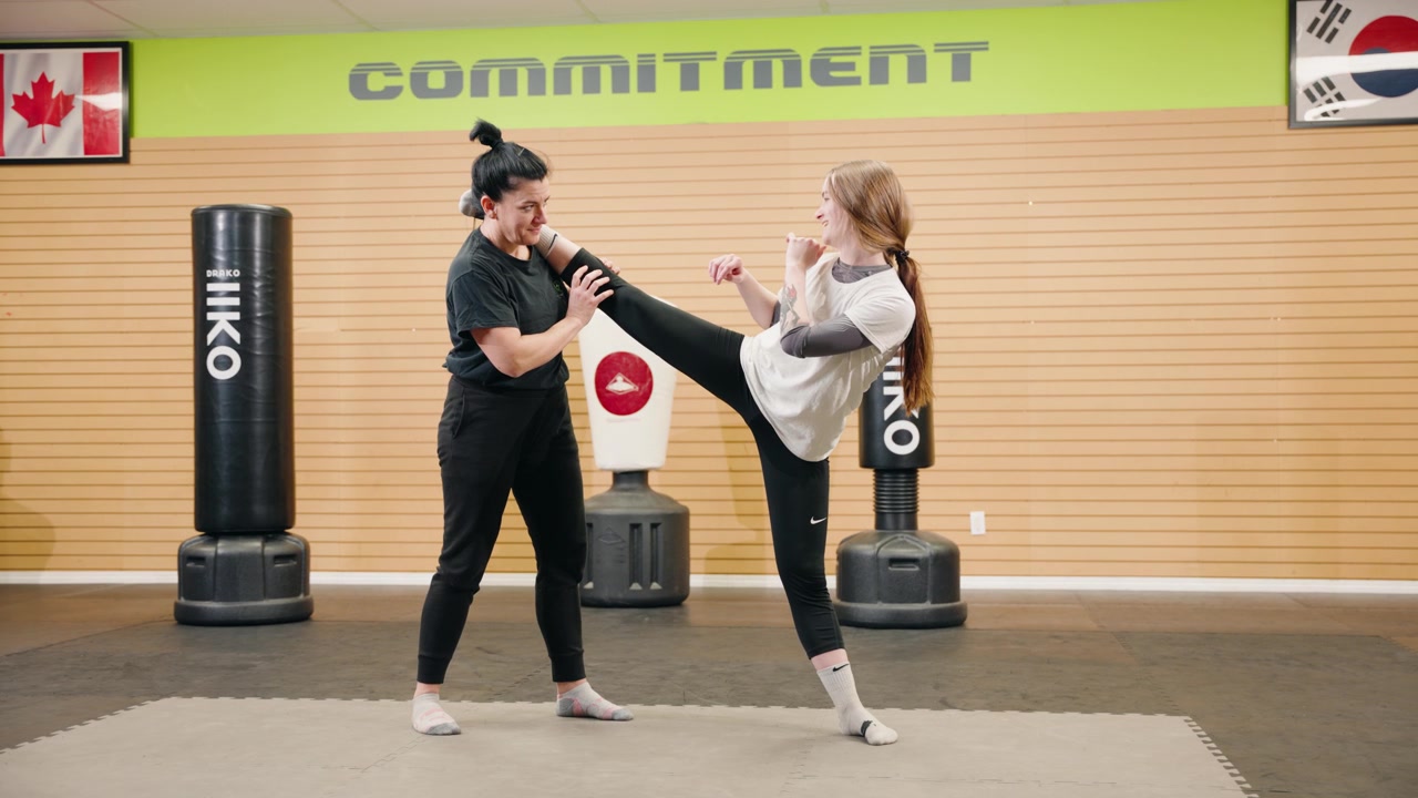 Bidikan Lebar: Ibu paruh baya, wanita, dan anak perempuan dalam kelompok atletis melakukan peregangan split berdiri di dalam pusat kebugaran gym seni bela diri Taekwondo, dengan latar belakang samsak, menunjukkan komitmen, serta bendera Korea dan Kanada.