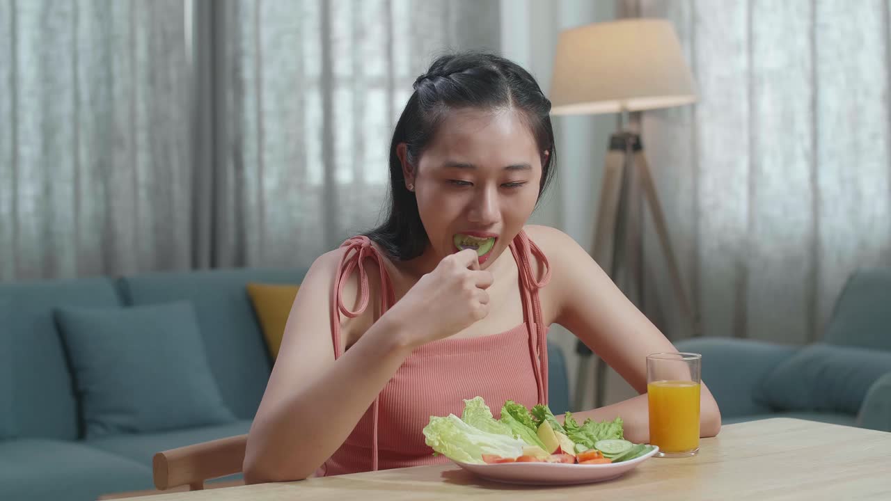 건강한 음식과 오렌지 주스 한 잔을 가진 미소 짓는 아시아 여성이 집에서 먹기 전에 오징어를 르는 포크를 사용합니다.