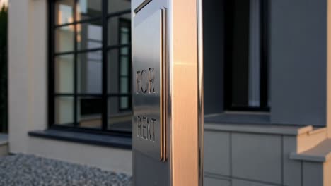 metal for rent sign reflecting golden sunset light standing in front of modern house with large windows and gravel, concept of real estate market and housing availability