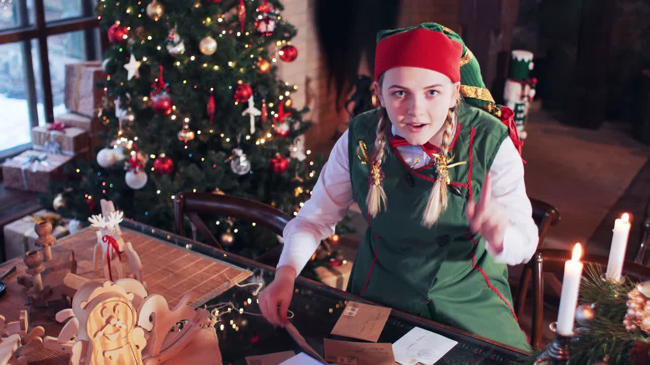 Merry elf girl in a green suit transfers letters on a table in a room with a Christmas tree and decorations. Against the background of the fireplace