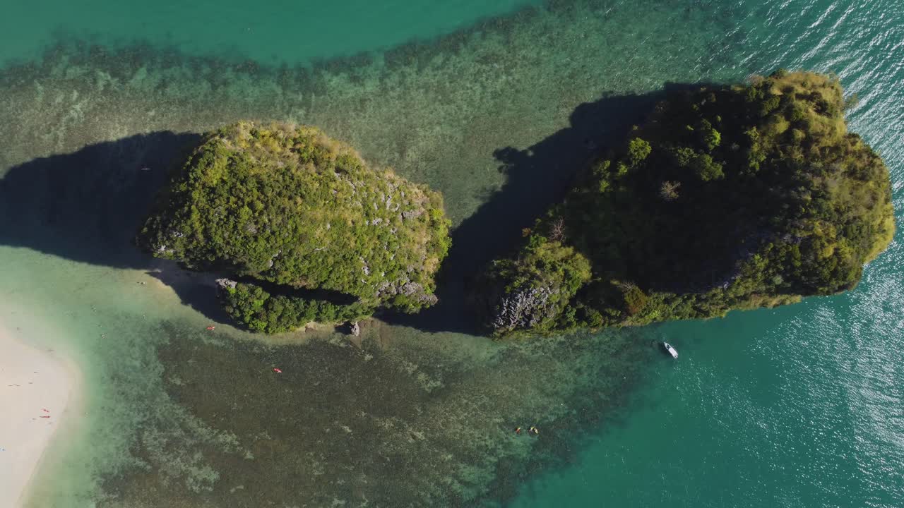 Top-down view of two small green islands off the Ao Nang coast, surrounded by coral reefs and shallow turquoise water, with visible kayakers and beachgoers on the nearby shore.