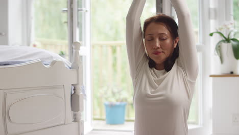 mature asian woman in pyjamas sitting on bedroom floor meditating in yoga pose - shot in slow motion