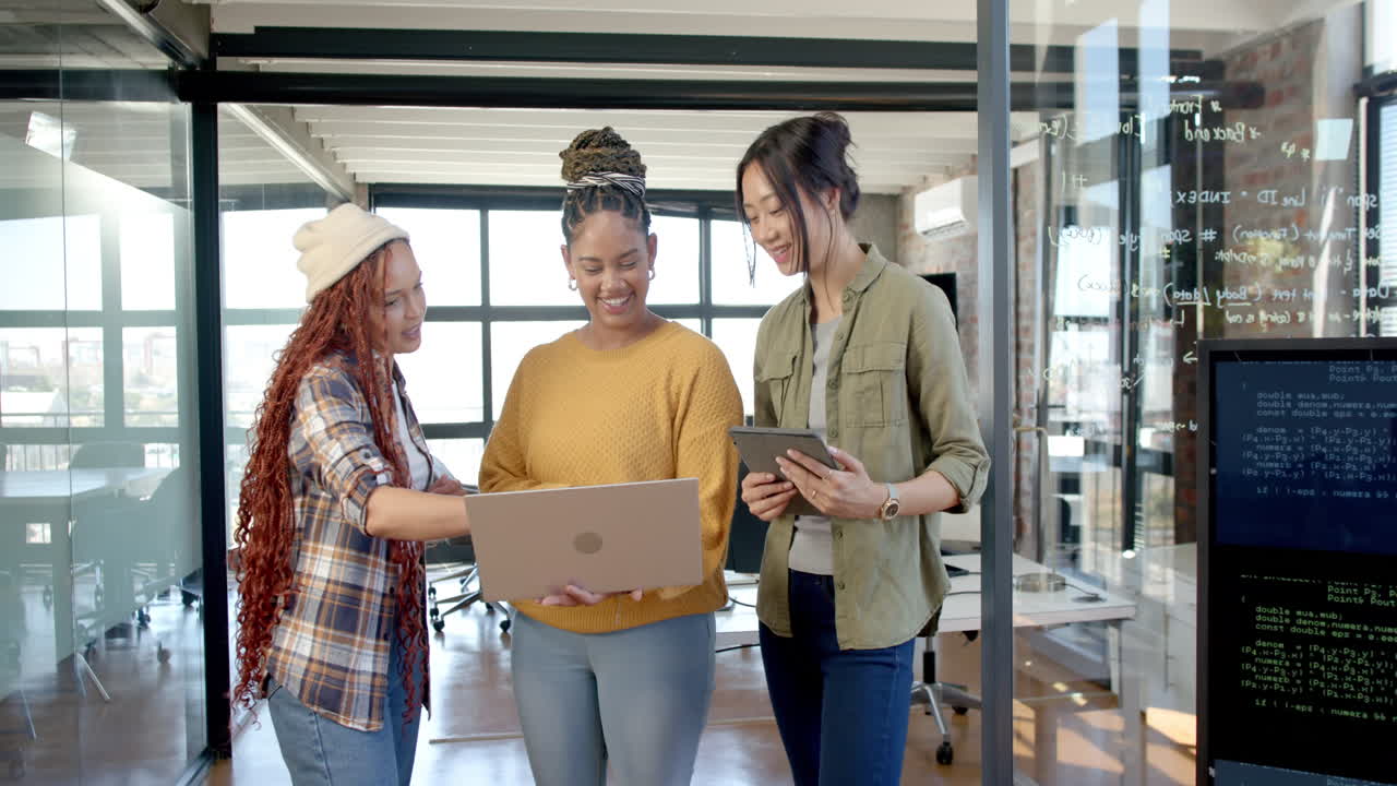 Diverse female team collaborating on laptop and tablet in modern office, discussing code