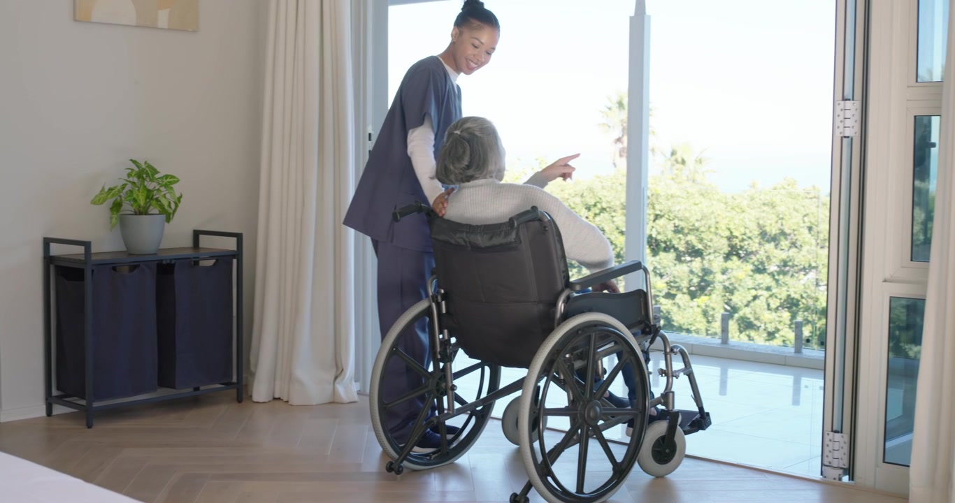紺色のスクラブを着たアフリカ系の介護士が、車椅子の高齢のアジア人をバルコニーへ案内し、景色を見せている。