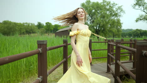 young woman in sundress walks towards camera on boardwalk through tall grass