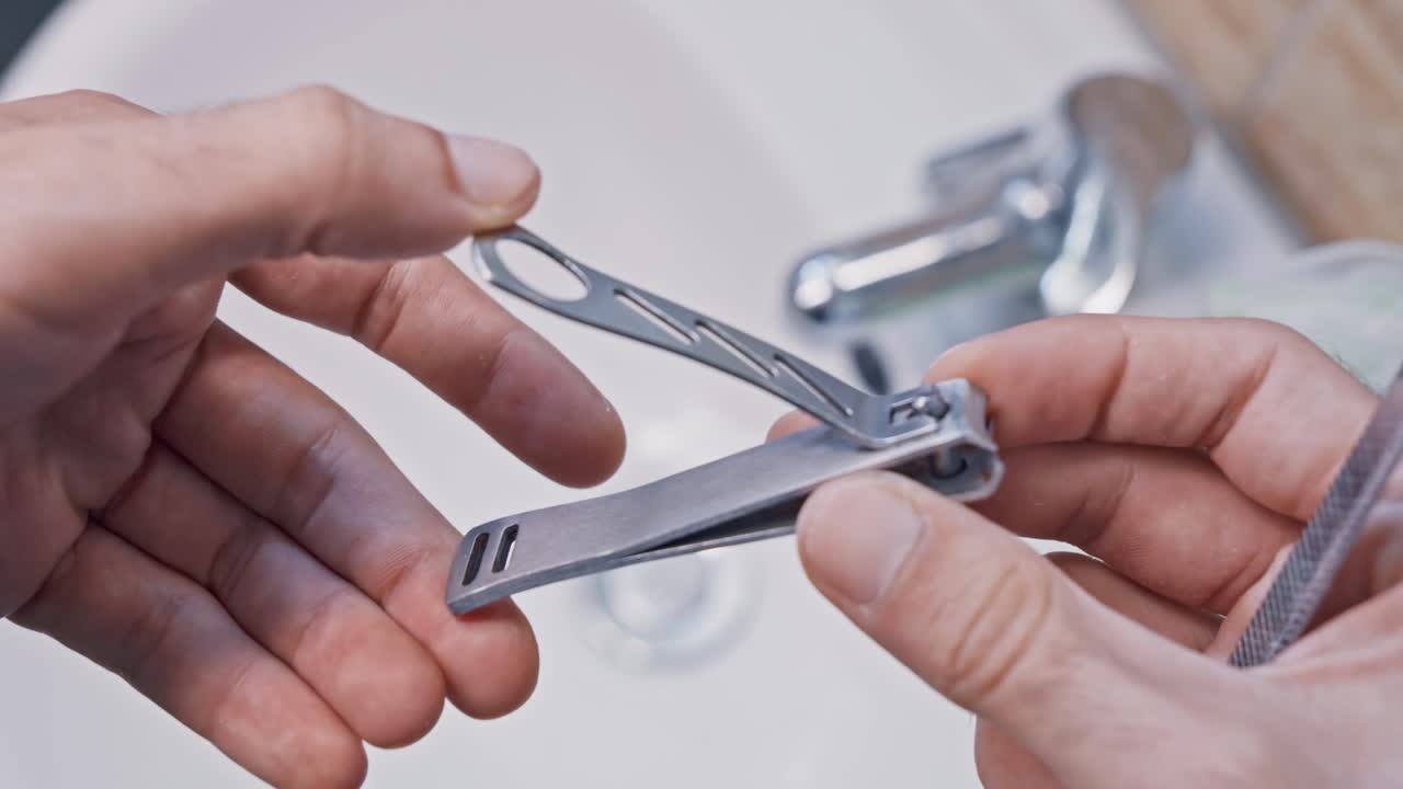 Close up of male hands holding manicure tools. A person is showing a nail clipper and a file in a bathroom over a white ceramic sink
