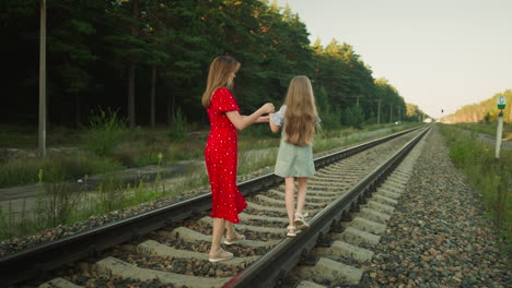 rear view of woman in red dress gently holding little girl hand for balance while walking along railway track surrounded by forest trees and gravel path under soft natural daylight