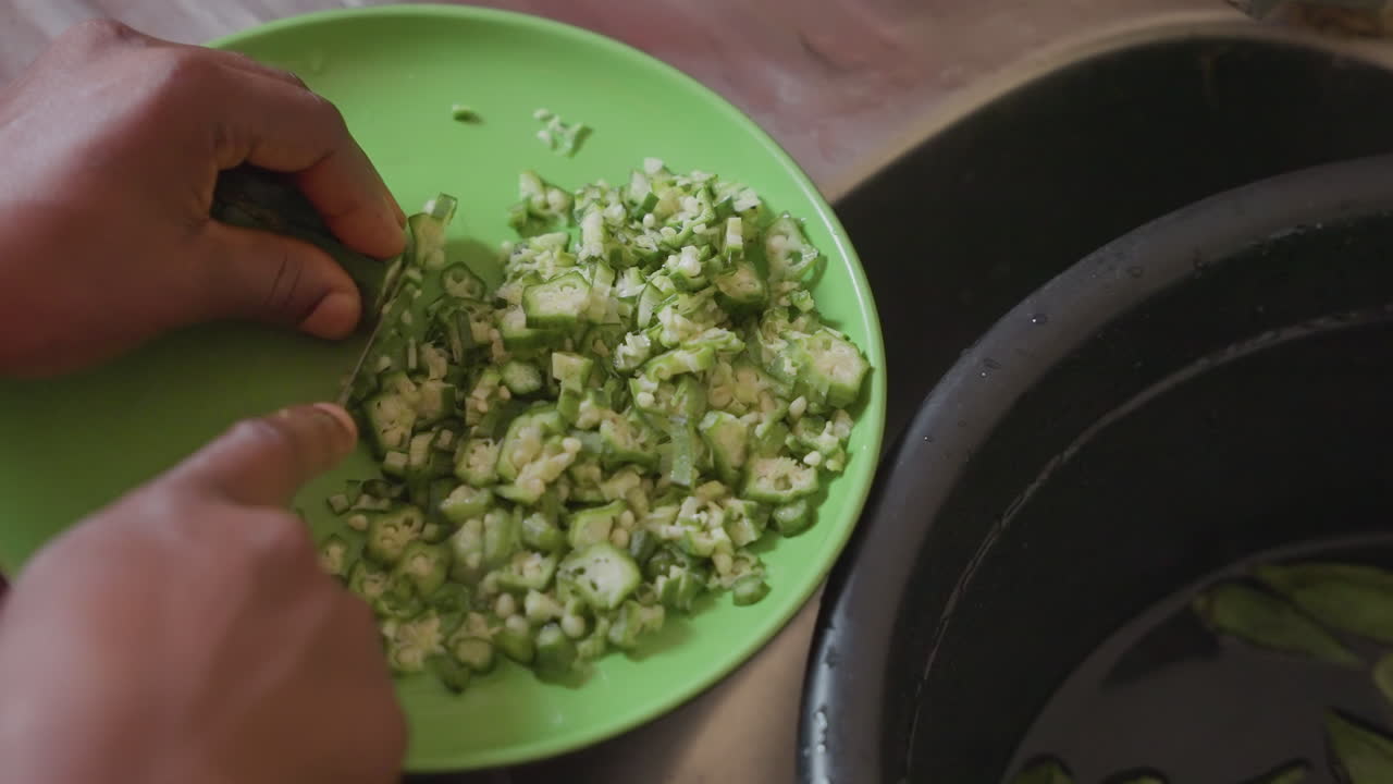 Black hands chopping green vegetable in plastic bowl beside sink home cook prepping fresh salad pieces with small knife, closeup tactile focus, natural light on damp basin, textured chopped