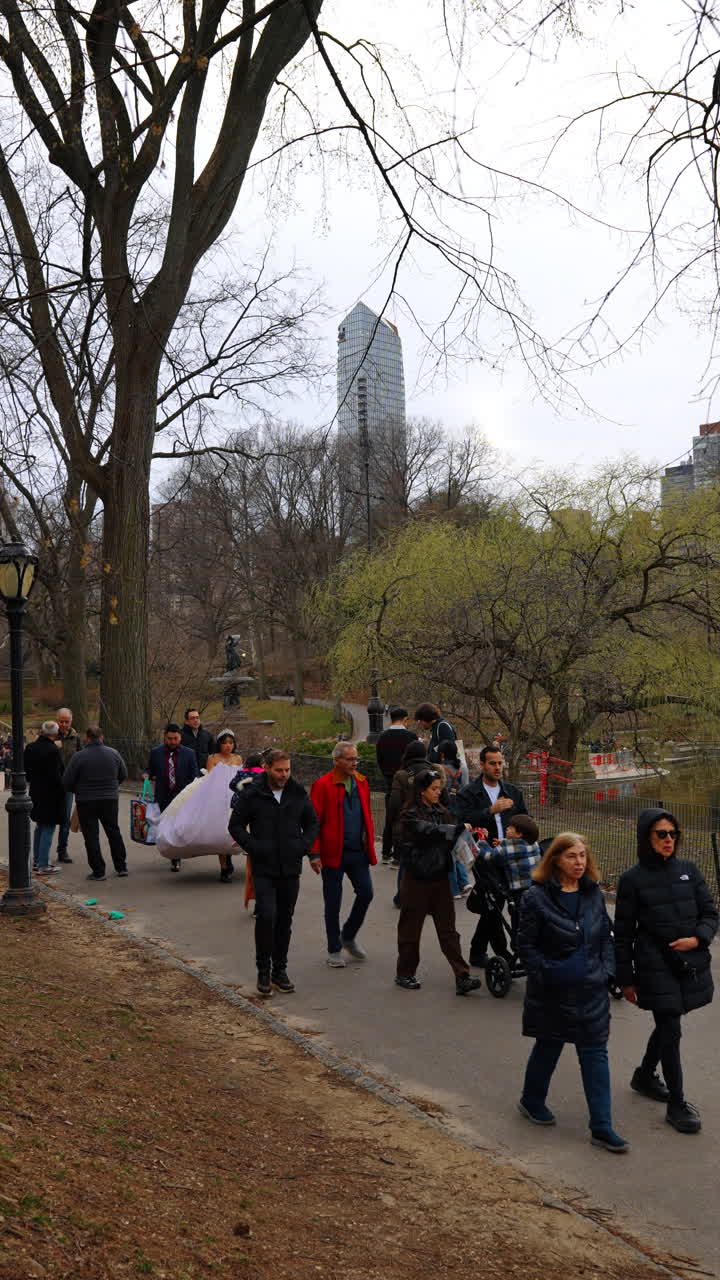 New York, USA, 14 marzo 2026: Persone che camminano su un sentiero pedonale a Central Park. Gruppo di persone che camminano su un sentiero lastricato in un parco con alberi spogli e un grattacielo in lontananza