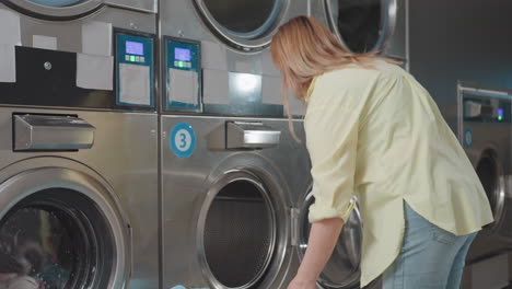 Laundry woman carries basket of clothes, walks toward industrial washer, door open, prepares loading garments into drum inside laundromat, stainless panels, chores, hygiene, service in operation
