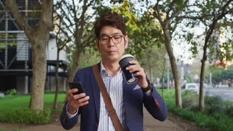 hombre asiático sosteniendo una taza de café usando un teléfono inteligente mientras camina en el parque