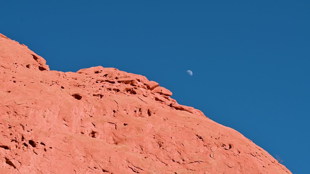 Aerial shot of moon rising over sunlit red rock ridge in Garden of the Gods, Colorado, with clear blue sky backdrop