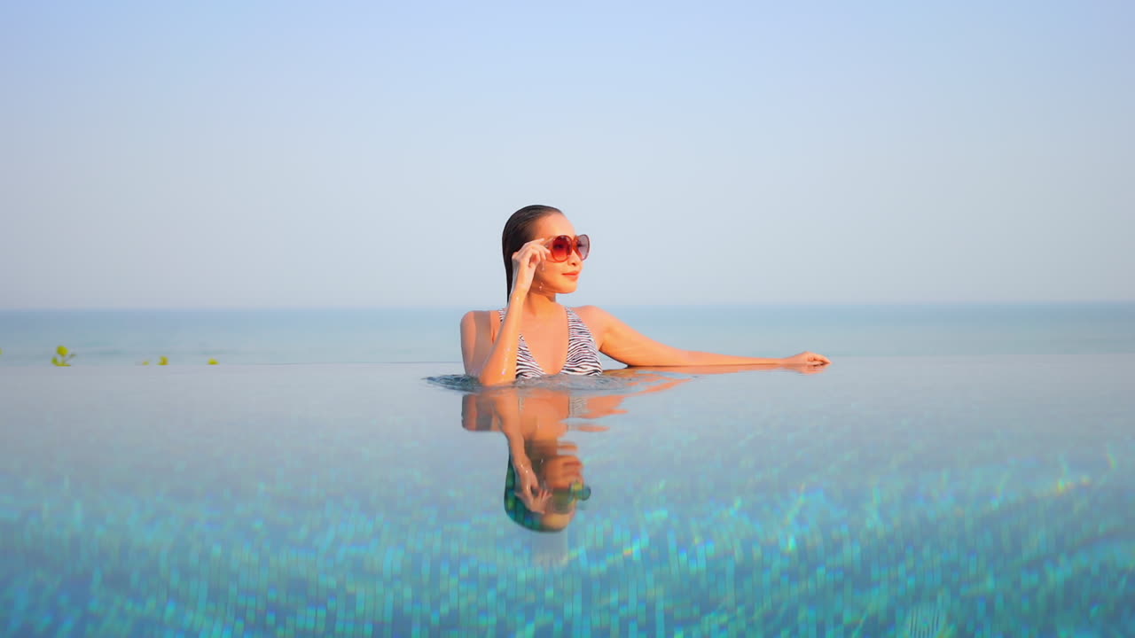 Junge asiatische Frau mit Sonnenbrille entspannt sich in einem Infinity-Pool mit türkisfarbenem Meer im Hintergrund