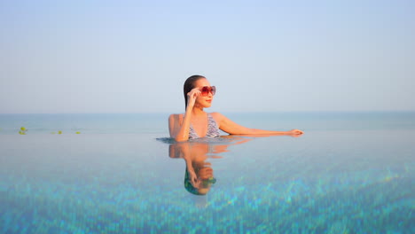 Joven-Asiática-Con-Gafas-De-Sol-Relajándose-En-La-Piscina-Infinita-Y-El-Mar-Turquesa-De-Fondo