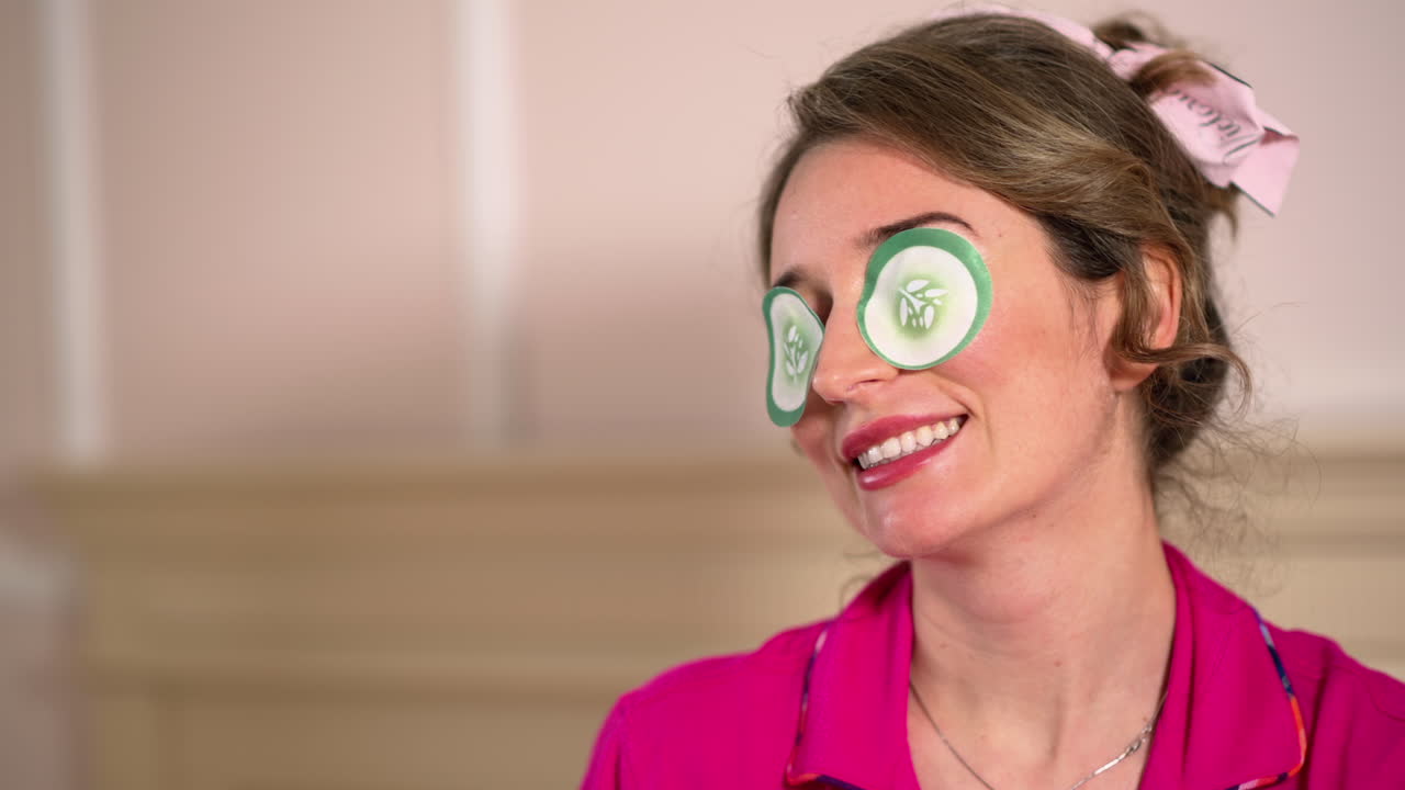 Brunette woman in pink pyjamas wearing eyepatches in front of a mirror, on the bed