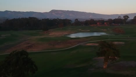 Vista-Aérea-Del-Campo-De-Golf-Con-Las-Montañas-Y-El-Lago-Al-Fondo