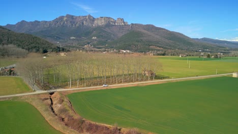 drone tracking a white car on a narrow road in an agricultural landscape with cultivated green fields and mountains in the background