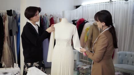 Two-caucasian-female-tailors-collaborating-on-white-wedding-dress-and-pinning-unfinished-garment-on-mannequin-in-fashion-studio,-fixing-sleeves.-Clothes-and-hangers-on-blurred-background